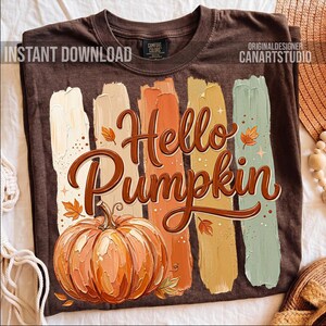 Puede incluir: Camiseta marr&oacute;n con un dise&ntilde;o tem&aacute;tico de oto&ntilde;o. El dise&ntilde;o presenta las palabras "Hello Pumpkin" en una fuente decorativa, una ilustraci&oacute;n de calabaza y pinceladas coloridas. La camiseta tambi&eacute;n tiene el texto "INSTANT DOWNLOAD" y "ORIGINALDESIGNER CANARTSTUDIO".