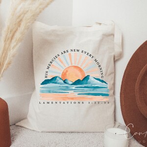 Op de afbeelding: Een witte canvas tote bag met een aquarel zonsopgang ontwerp en de tekst "His mercies are new every morning" en "Lamentations 3:22-33".