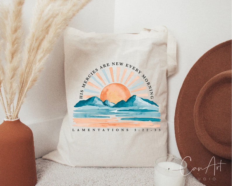 Op de afbeelding: Een witte canvas tote bag met een aquarel zonsopgang ontwerp en de tekst "His mercies are new every morning" en "Lamentations 3:22-33".