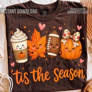 May include: A dark brown t-shirt featuring a graphic of a coffee cup, an orange maple leaf, a football, and a pumpkin, all with smiling faces. The text 'tis the Season' is printed in orange below the characters, accented by small pink hearts.