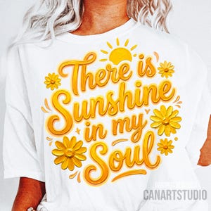 Peut inclure: T-shirt blanc avec l'inscription "There is Sunshine in my Soul" en jaune. Le motif comprend un soleil jaune et des fleurs jaunes. Les mots "Original Designer" et "CANARTSTUDIO" sont imprim&eacute;s en bas.