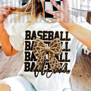 May include: Cream-colored t-shirt with the words "BASEBALL MOM" in black block letters. A large, tan and brown leopard print bow is centered on the shirt. The shirt is a casual, everyday style.