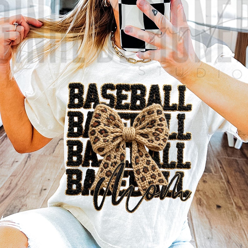 May include: Cream-colored t-shirt with the words "BASEBALL MOM" in black block letters. A large, tan and brown leopard print bow is centered on the shirt. The shirt is a casual, everyday style.