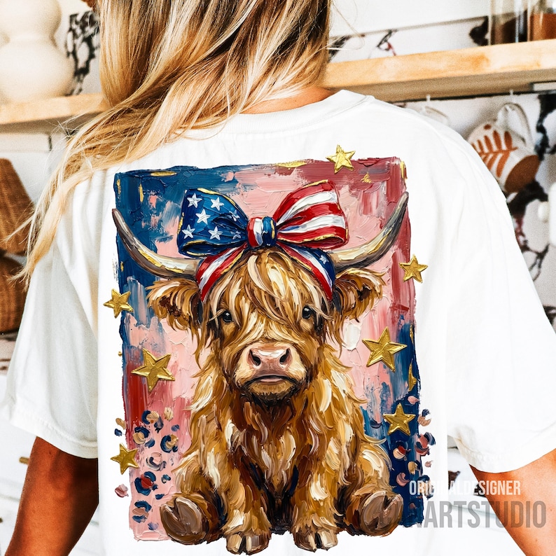 Peut inclure: T-shirt blanc avec un motif de vache des Highlands orn&eacute; d'un n&oelig;ud aux couleurs du drapeau am&eacute;ricain et d'&eacute;toiles dor&eacute;es. La vache a de longs poils bruns et hirsutes, sur un fond de coups de pinceau rouges, blancs et bleus. Le t-shirt porte le texte "ORIGINAL DESIGNER ARTSTUDIO".