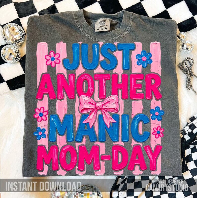 Op de afbeelding: Een grijs Comfort Colors T-shirt met de tekst "JUST ANOTHER MANIC MOM-DAY" in roze en blauw, versierd met roze bloemen en een strik. Het shirt ligt op een zwart-wit geblokt oppervlak.