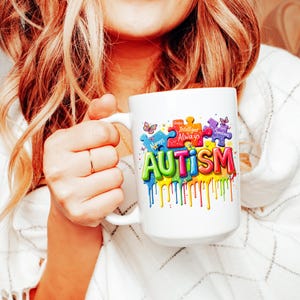 May include: White ceramic coffee mug with a colorful graphic of the word "AUTISM" with puzzle pieces and butterflies. The puzzle pieces have text that reads "Unique, Intelligent, Always, Sometimes".