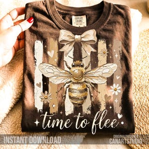Puede incluir: Camiseta marrón con una ilustración detallada de una abeja, lazo, flores y corazones. El texto "time to flee" está escrito en una fuente de escritura. La camiseta está doblada y sostenida por una mano.