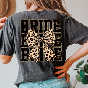 May include: A black t-shirt with the text "Bride Tribe" in gold glitter lettering. A large leopard print bow is centred over the text.