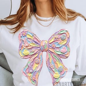 May include: White t-shirt featuring a large, textured bow design in pastel colors. The bow is decorated with Easter-themed elements like eggs, bunnies, and chicks. The text "ORIGINAL DESIGNER SANARTS STUDIO" is printed on the bottom right.