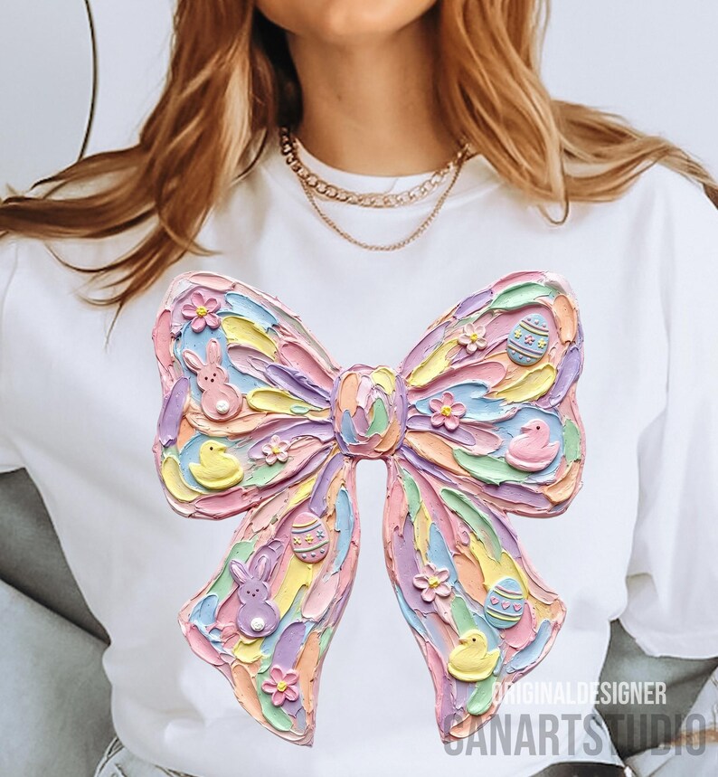 May include: White t-shirt featuring a large, textured bow design in pastel colors. The bow is decorated with Easter-themed elements like eggs, bunnies, and chicks. The text "ORIGINAL DESIGNER SANARTS STUDIO" is printed on the bottom right.