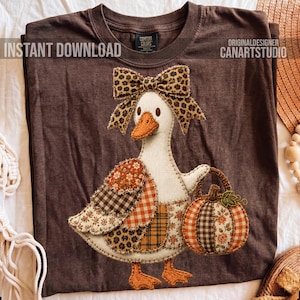 May include: A dark brown t-shirt featuring a graphic of a white goose with an orange beak. The goose wears a leopard print bow and carries a pumpkin made of various fabric patterns including floral, plaid, and gingham in orange and brown tones.