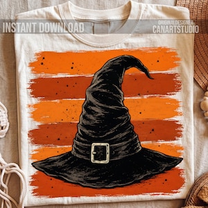 May include: A cream-coloured t-shirt featuring a graphic of a black witch hat with a buckle, centred over a background of horizontal orange and dark orange brushstrokes. The text INSTANT DOWNLOAD and CANARTSTUDIO appear at the top corners.
