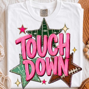 May include: A white t-shirt featuring a graphic design with the text TOUCH DOWN in bright pink letters. The design includes three stars: one green with football field markings, one green with play diagrams, and one brown with football laces.