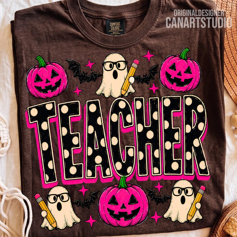 May include: A brown t-shirt featuring a graphic print with the word TEACHER in large black letters with white polka dots and a pink outline. The design includes three pink jack-o'-lanterns, three white ghosts wearing glasses, and yellow pencils.