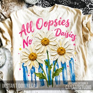 May include: Cream-colored t-shirt with the phrase "All Oopsies No Daisies" in pink script. The shirt features a floral design of three daisies with yellow centers and white petals, along with blue and white stripes and green leaves.