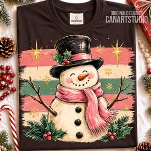 Może przedstawiać: Ciemnobrązowy t-shirt z grafiką bałwana w czarnym cylindrze i r&oacute;żowym szaliku. Bałwan ma marchewkowy nos, gałązkowe ramiona i trzy czarne guziki. Tło zawiera poziome paski, złote gwiazdki oraz gałązki ostrokrzewu z czerwonymi owocami.