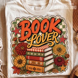 May include: A cream-colored t-shirt featuring a graphic with the text BOOK LOVER in orange and yellow. The design includes a stack of five books, colorful chrysanthemums, autumn leaves, red and yellow stars, and a green and white checkered background.