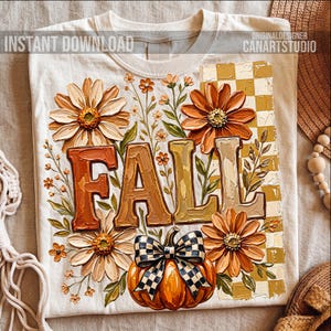May include: Cream-colored t-shirt with a fall-themed design. The design features the word "FALL" in large, textured letters, surrounded by orange and cream-colored flowers, a pumpkin with a checkered bow, and a checkered background.