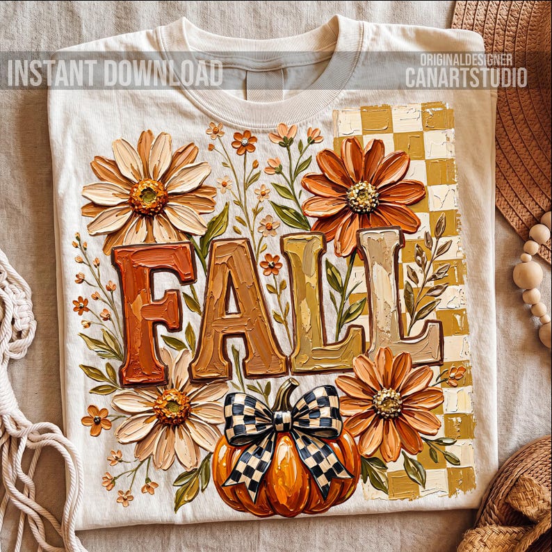 May include: Cream-colored t-shirt with a fall-themed design. The design features the word "FALL" in large, textured letters, surrounded by orange and cream-colored flowers, a pumpkin with a checkered bow, and a checkered background.