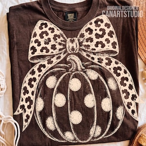 May include: A dark brown t-shirt featuring a large, cream-colored graphic of a pumpkin decorated with polka dots. A prominent bow with a leopard print pattern sits atop the pumpkin. The text ORIGINALDESIGNER CANARTSTUDIO is printed in the corner.