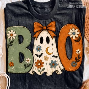 Può includere: Una t-shirt grigio scuro con la scritta BOO in grandi lettere. La B è verde salvia con fiori bianchi, la O è un fantasma bianco con un fiocco arancione con fiori, stelle, una luna e pipistrelli, e la O finale è arancione con fiori bianchi e blu.
