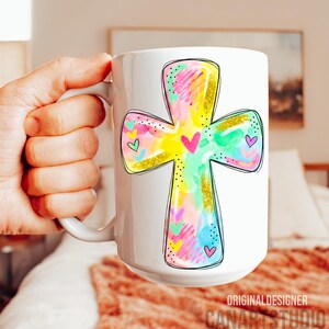 Op de afbeelding: Witte keramische mok met een kleurrijk kruisontwerp met zwarte omtrek. Het kruis is gevuld met pastelkleuren, goudglitter en hartaccenten. De mok heeft onderaan de tekst "ORIGINAL DESIGNER CANARTSTUDIO".