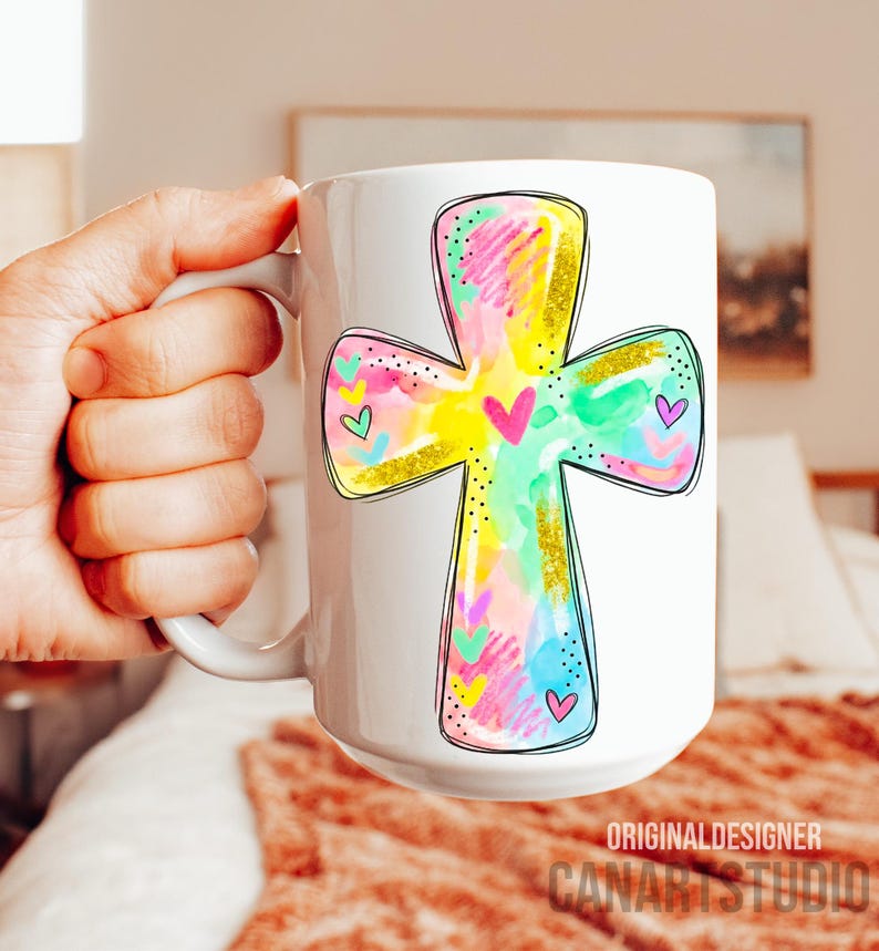 Op de afbeelding: Witte keramische mok met een kleurrijk kruisontwerp met zwarte omtrek. Het kruis is gevuld met pastelkleuren, goudglitter en hartaccenten. De mok heeft onderaan de tekst "ORIGINAL DESIGNER CANARTSTUDIO".