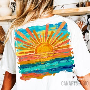 May include: White t-shirt featuring a vibrant, impressionistic painting of a sunset. The artwork depicts a bright yellow sun with orange and yellow rays over a colorful horizon of orange, pink, and blue. The text "CANARTSTUDIO" is printed on the bottom right.