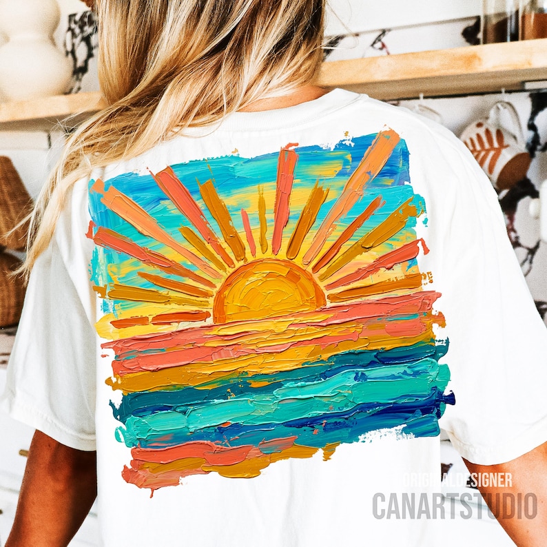 May include: White t-shirt featuring a vibrant, impressionistic painting of a sunset. The artwork depicts a bright yellow sun with orange and yellow rays over a colorful horizon of orange, pink, and blue. The text "CANARTSTUDIO" is printed on the bottom right.