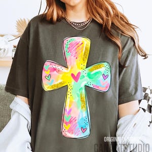 Op de afbeelding: Donkergrijs t-shirt met een kleurrijk kruisontwerp. Het kruis is versierd met aquarelspatten in roze, geel, groen en blauw, geaccentueerd met gouden glitter en hartdetails. De tekst "ORIGINAL DESIGNER CANA STUDIO" staat rechtsonder.