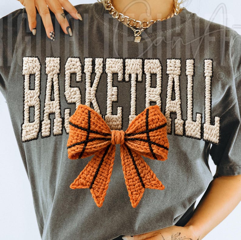 Op de afbeelding: Een grijs t-shirt met het woord "BASKETBALL" in een cr&egrave;mekleurig, getextureerd lettertype. Een gehaakte oranje strik, ontworpen om op een basketbal te lijken, is gecentreerd onder de tekst. De strik heeft zwarte lijnen die het ontwerp van de basketbal nabootsen.