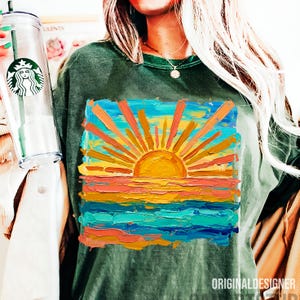 May include: Dark green t-shirt featuring a vibrant, impressionistic painting of a sunset over the ocean. The artwork uses bold brushstrokes and a palette of orange, yellow, blue, and teal. The text "ORIGINALDESIGNER" is printed at the bottom.