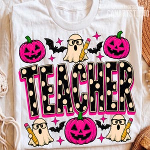 May include: A white t-shirt featuring a graphic print with the word TEACHER in large black letters with white polka dots. The design includes three pink jack-o'-lanterns, three ghosts wearing black glasses and holding yellow pencils, and black bats.