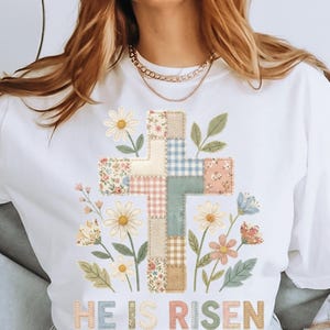May include: White t-shirt featuring a patchwork cross design with floral and gingham patterns, surrounded by daisies and other flowers. The text "HE IS RISEN" is stitched at the bottom, creating a spring or Easter theme.