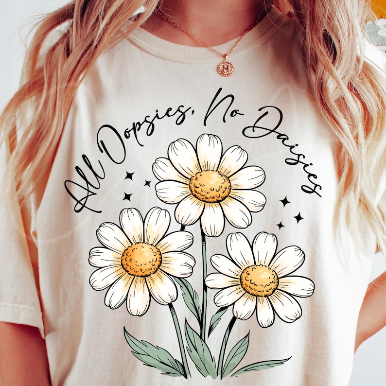 May include: Cream-colored t-shirt featuring a daisy graphic with the text "All Oopsies, No Daisies." The design includes three large white daisies with yellow centers and green stems and leaves. Small black star accents surround the flowers.
