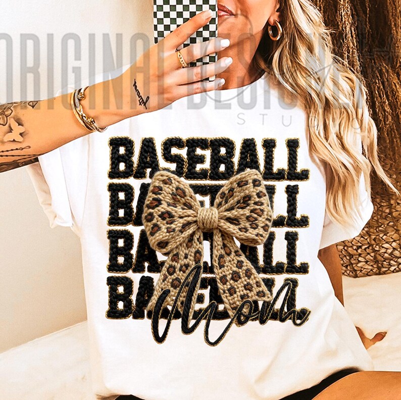 May include: White t-shirt with the words "BASEBALL MOM" in black and gold lettering. A tan and brown leopard print bow is centered on the shirt. The shirt is a casual style.