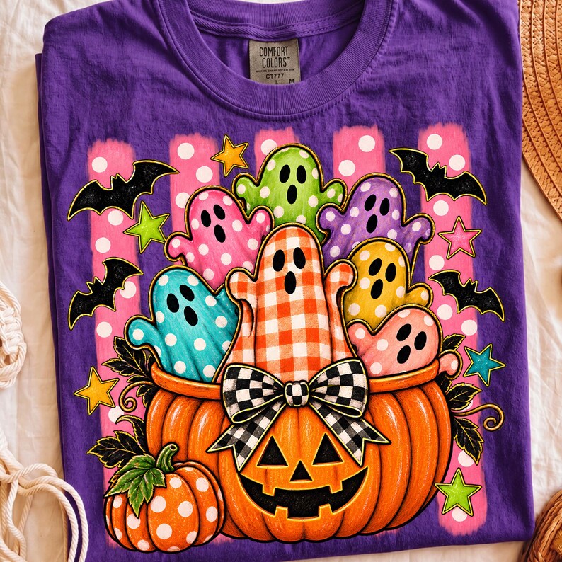 May include: A purple t-shirt with a graphic print of a large orange jack-o'-lantern filled with colorful ghosts in polka dot and gingham patterns. The design includes black bats, small stars, and a black and white checkered bow on the pumpkin.