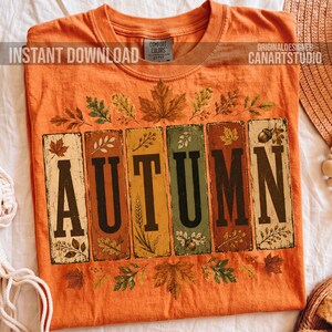 May include: An orange t-shirt featuring the word AUTUMN in large black letters, with each letter enclosed in a rectangular panel. The design includes illustrations of various autumn leaves, wheat stalks, and acorns surrounding the text panels.