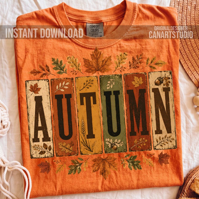 May include: An orange t-shirt featuring the word AUTUMN in large black letters, with each letter enclosed in a rectangular panel. The design includes illustrations of various autumn leaves, wheat stalks, and acorns surrounding the text panels.