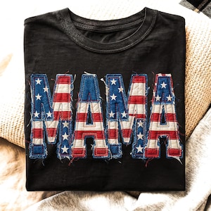 May include: Black t-shirt with the word "MAMA" in a patriotic design. The letters are made to look like the American flag, with blue star-filled sections and red and white stripes. The shirt has a distressed look.