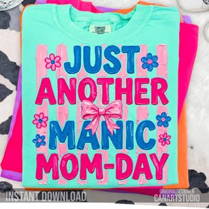 Op de afbeelding: Een turquoise Comfort Colors t-shirt met de tekst "JUST ANOTHER MANIC MOM-DAY" in roze en blauw, versierd met bloemen en een strik. Het shirt is opgevouwen bovenop andere felgekleurde shirts.