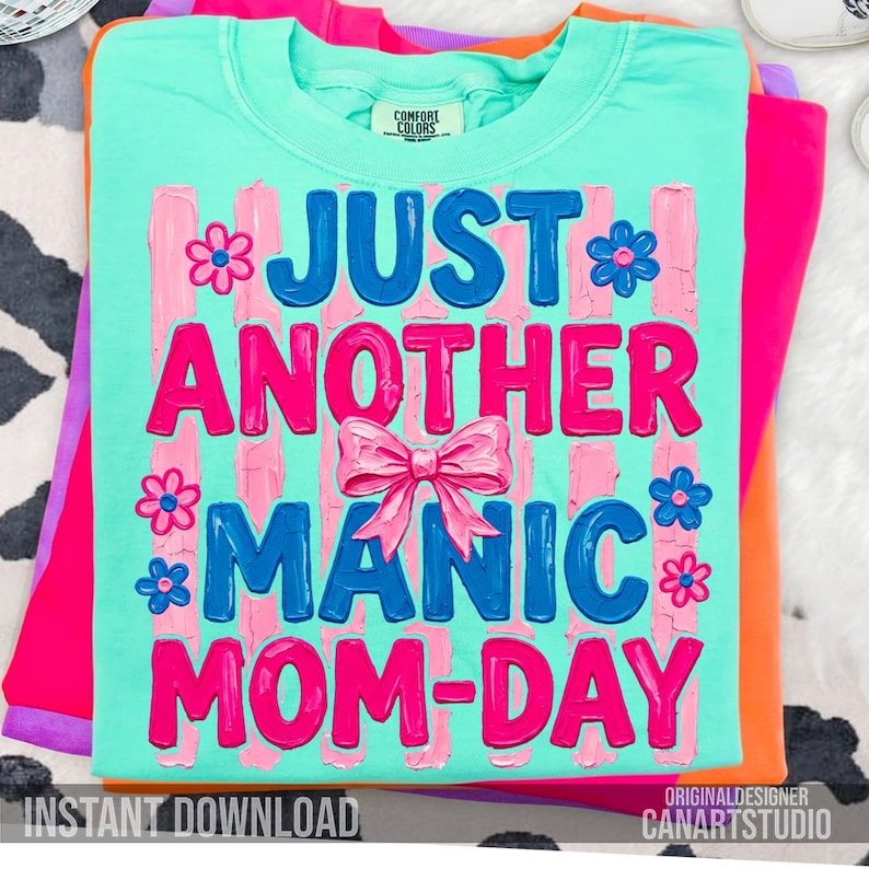 Op de afbeelding: Een turquoise Comfort Colors t-shirt met de tekst "JUST ANOTHER MANIC MOM-DAY" in roze en blauw, versierd met bloemen en een strik. Het shirt is opgevouwen bovenop andere felgekleurde shirts.