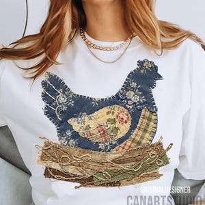 May include: White t-shirt featuring a patchwork hen design in blue floral, yellow floral, and plaid fabrics. The hen sits on a nest made of brown and green fabric strips. The text "ORIGINAL DESIGNER CANARTS STUDIO" is printed at the bottom.