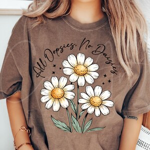 May include: Brown t-shirt featuring a floral graphic of three white daisies with yellow centers and green stems. The text "All Dopsies, No Daisies" is arched above the flowers in a handwritten style.