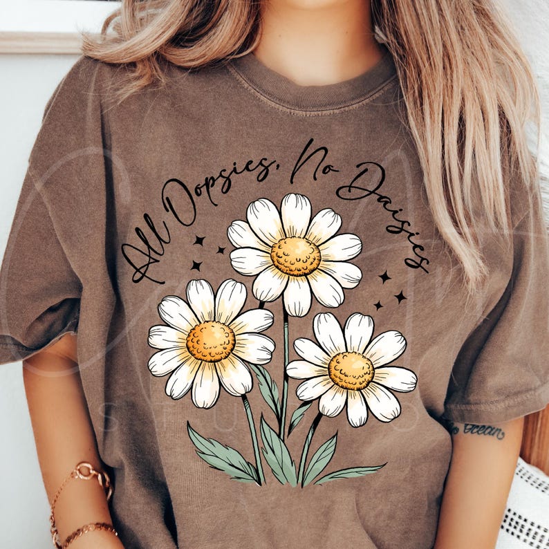 May include: Brown t-shirt featuring a floral graphic of three white daisies with yellow centers and green stems. The text "All Dopsies, No Daisies" is arched above the flowers in a handwritten style.