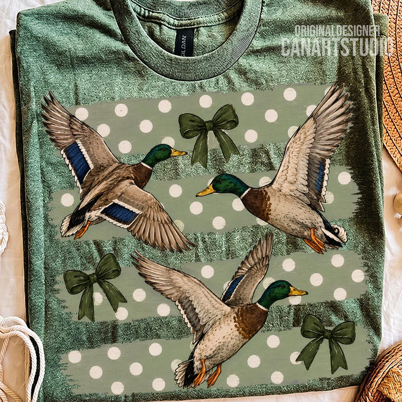 Pu&ograve; includere: Una t-shirt verde oliva m&eacute;lange con una stampa grafica di tre germani reali in volo. Il design presenta bande orizzontali verde chiaro a pois bianchi e tre fiocchi verde scuro. &Egrave; presente il testo ORIGINAL DESIGNER CANARTSTUDIO.