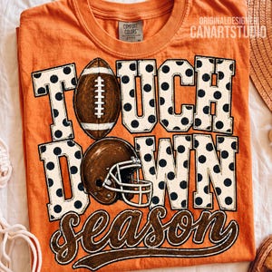 May include: An orange crew neck t-shirt featuring the text TOUCH DOWN SEASON in large white block letters with black polka dots. A brown football replaces the letter O in TOUCH, and a brown football helmet is positioned over the letter O in DOWN.