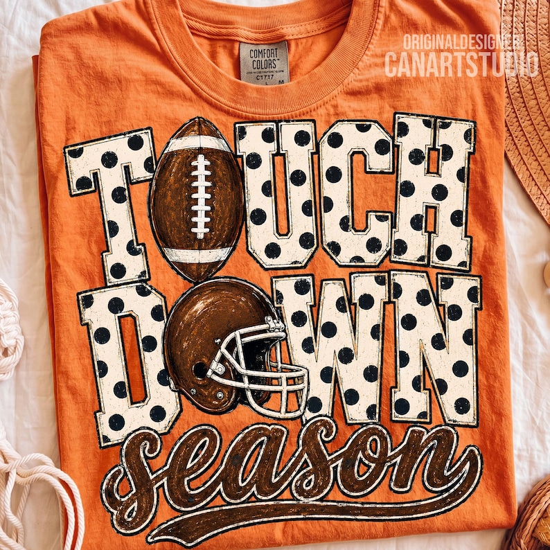 May include: An orange crew neck t-shirt featuring the text TOUCH DOWN SEASON in large white block letters with black polka dots. A brown football replaces the letter O in TOUCH, and a brown football helmet is positioned over the letter O in DOWN.