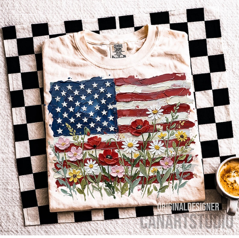 May include: Cream-colored t-shirt featuring a textured American flag design with red and white stripes, blue stars, and colorful flowers. The shirt is folded on a black and white checkered cloth.