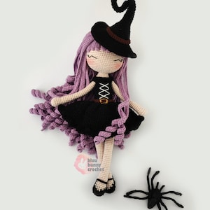 May include: A crocheted witch doll with purple hair, a black dress, and a black witch hat. The doll is lying on its back with its legs crossed. A black crocheted spider is on the white surface below the doll. The doll's eyes are closed and it has a sweet expression. The doll is wearing a black belt with a buckle. The doll's hair is in a curly style. The doll is made by @bluubunnycrochet.
